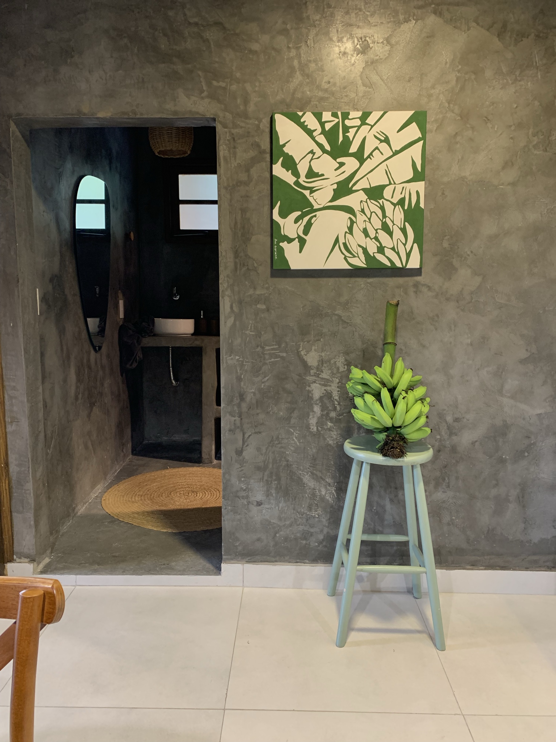 Parede de cimento com quadro de folhas em verde e branco e banco com cacho de bananas